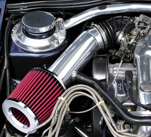 Roter, offener Sportluftfilter im Motorraum, verbunden mit einem glänzend silbernen Cold Air Intake Rohr zum Motorblock. Umgeben von silbernen Motorteilen, schwarzen Schläuchen und geflochtenen Leitungen sticht der Filter hervor.